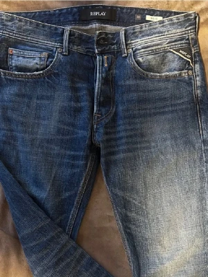 Blå Replay jeans med slitningar - Snygga blå jeans från Replay med klassisk femficksmodell och tydliga slitningar framtill. Jeansen har en slim fit-passform, detaljerade sömmar på bakfickorna och läderpatch med Replay-logga bak i midjan. Tillverkade i bomull med en cool tvättad look. Knappen är av, men går lätt att fixa!