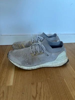 Adidas Ultra Boost grå sneakers - Grå Adidas Ultra Boost sneakers med stickad ovandel och vita Boost-sulor. Skorna har lågt skaft, snörning och en strumpliknande passform. Diskreta detaljer i beige och svart på sulan. Perfekta för dig som gillar sportig och clean stil.