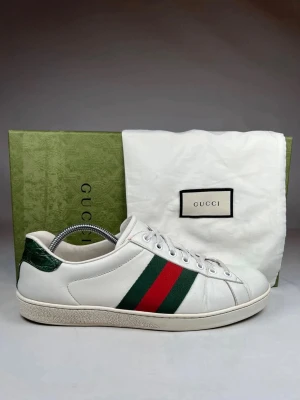 Gucci Ace sneakers  - Klassiska vita sneakers från Gucci i läder med gröna och röda ränder på sidan samt grön detalj vid hälen. Snygg och tidlös design med lågt skaft och vita snören. Perfekta för dig som vill ha en ikonisk och stilren sko.