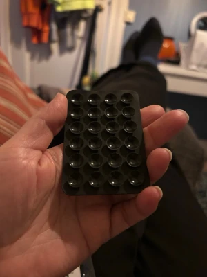 Svart mobilhållare med sugproppar - Praktisk svart mobilhållare med flera små sugproppar på ena sidan och självhäftande baksida. Perfekt för att fästa mobilen på olika ytor, t.ex. i bilen eller på skrivbordet. Tillverkad i plast och silikon för bra grepp och flexibilitet.