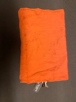 Orange halsduk från No Name Sweden - Snygg orange halsduk från No Name Sweden i mjukt och tunt tyg. Halsduken har fransade kanter och en enkel, stilren design som passar perfekt till höstens outfits. Materialet känns lätt och luftigt, vilket gör den enkel att bära och styla.