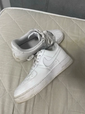 Nike Air Force 1  - Klassiska vita Nike Air Force 1 sneakers med lågt skaft och ikonisk swoosh på sidan. Skorna är i helvitt läder med perforerad tå för ventilation och har en tjock sula med AIR-dämpning. Perfekta för en clean och stilren look. Lite rengöring så är den på topp