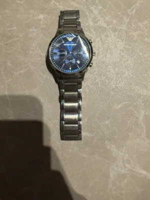 Emporio Armani blå klocka i stål - Snygg klocka från Emporio Armani med blå urtavla och silverfärgad länk i rostfritt stål. Klockan har flera små urtavlor och datumvisning. Perfekt accessoar för dig som vill ha en stilren och modern look.