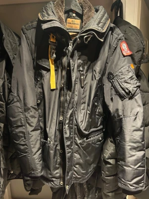 Svart dunjacka från Parajumpers - Snygg svart dunjacka från Parajumpers med flera fickor, patch på ärmen och gul detalj vid kragen. Jackan har hög krage, dragkedja och knappar samt fodrad insida för extra värme. Perfekt för kalla vinterdagar.