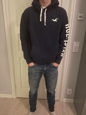 Svart hoodie från Hollister - Svart hoodie från Hollister med vit logga på bröstet och stort vitt tryck längs ena ärmen. Klassisk känguruficka framtill och vita dragsnören i huvan. Perfekt för en avslappnad och trendig stil.