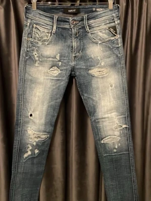 Replay slitna blå jeans herr - Säljer ett par blå slim fit jeans från Replay med slitningar, hål och snyggt tvättad look. Jeansen har fem fickor, dragkedja och knappgylf samt Replay-logga på fickan. Perfekta för en avslappnad och trendig stil.