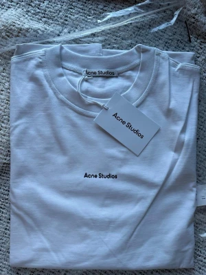 Vit t-shirt från Acne S. - Vit t-shirt från Acne S. i mjuk bomull med minimalistisk logga tryckt på bröstet. Klassisk rund halsringning och korta ärmar. Perfekt för en clean och stilren look.
