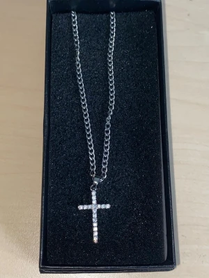 Silverfärgat halsband med korshänge 55cm - Snyggt silverfärgat halsband med ett korsformat hänge dekorerat med små glittrande stenar. Kedjan är av metall och har en klassisk länkdesign. Perfekt accessoar för dig som gillar stilrena och trendiga smycken.