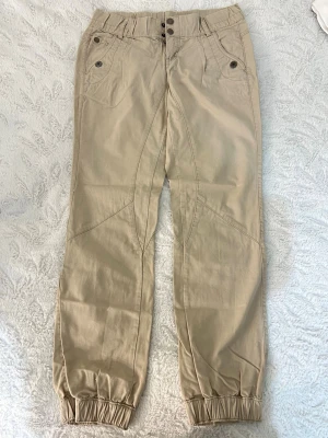 Beiga cargobyxor från Kappahl - Snygga beiga cargobyxor från KappAhl med elastisk midja och muddar vid benslut. Byxorna har flera fickor framtill och coola sömdetaljer. Skriv om flera bilder önskas 🥰