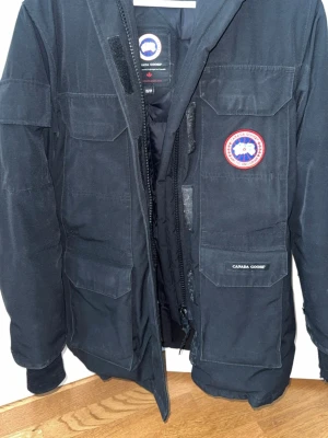 Svart dunjacka från Canada Goose XS - Säljer en svart dunjacka från Canada Goose med klassisk patch på bröstet och flera praktiska fickor framtill. Jackan har dragkedja och kardborrestängning samt hög krage för extra värme. Perfekt för kalla vinterdagar.
