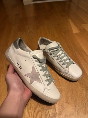 Vita Golden Goose sneakers med stjärna - Snygga vita sneakers från Golden Goose med grå snörning och en ikonisk grå stjärna på sidan. Skorna har en låg siluett och detaljer i silver vid hälen. Tillverkade i läder med klassisk rund tå och platt sula. Perfekta för en trendig och avslappnad stil. I princip nya och har endast vart använda ett fåtal gånger🙌