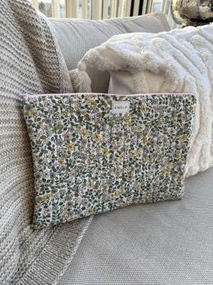 Blommig datorväska från Emelo - Säljer en datorväska från Emelo med blommigt mönster i grönt, gult och rosa på vit botten. Väskan är rektangulär med dragkedja upptill i ljusrosa och är tillverkad i bomull. Perfekt för att bära din laptop med stil. Vadderat. Passar laptop med maxmått: 33x24 cm