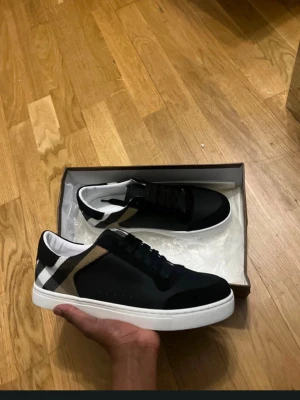Svarta Burberry sneakers med rutmönster - Snygga svarta sneakers från Burberry med klassiskt beige, vitt och svart rutmönster på hälen. Skorna har vita sulor, svarta snören och är tillverkade i en mix av läder och textil. Perfekta för dig som vill ha en stilren och trendig look.
