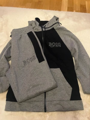 Grå hoodie från Hugo Boss med svart detalj - Snygg grå hoodie från Hugo Boss med svarta paneler och dragkedja framtill. Hoodien har huva med dragsko och logga på bröstet. Tillverkad i mjukt sweatshirtmaterial som är perfekt för chill dagar. Matchande byxor medföljer, båda med tydlig BOSS-logga.