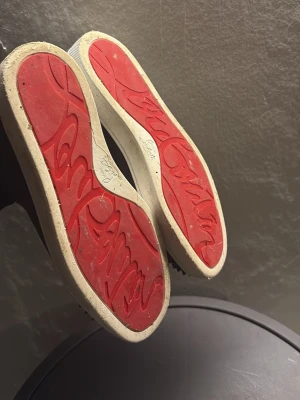 Christian Louboutin svarta sneakers - Svarta sneakers från Christian Louboutin med klassisk röd sula och coola nitar på tån. Skorna har snörning och är tillverkade i mocka med detaljer i läder. Perfekta för dig som vill sticka ut med en edgy och lyxig stil.