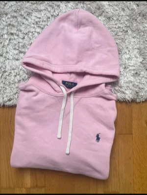 Ljusrosa hoodie från Polo Ralph Lauren - Säljer en ljusrosa hoodie från Polo Ralph Lauren med klassisk huva och vita dragsnören. Tröjan har en broderad marinblå logga på bröstet och en stor magficka framtill. Skriv vid minsta lilla fråga.