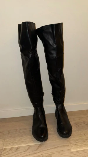 Svarta overknee stövlar  - Säljer ett par svarta overknee stövlar med chunky sula och dragkedja på insidan. Tillverkade i skinnimitation med matt finish. Stövlarna har rund tå och en hög, mjuk skaft som går över knät. Perfekta för en edgy och trendig look.