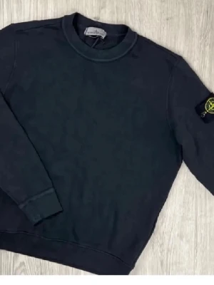 Svart sweatshirt från Stone Island - Svart sweatshirt från Stone Island med klassisk rund hals och ribbade muddar. Ikonisk logotyp-patch på vänster ärm. Perfekt för en stilren och avslappnad look.
