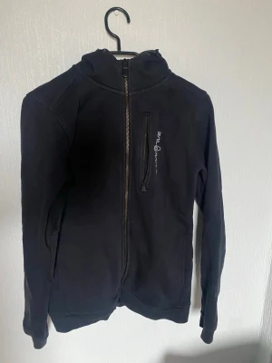 Svart hoodie från Sail Racing - Säljer denna svarta hoodie från Sail Racing med dragkedja och huva. Perfekt för en casual look. Den har en logotyp på bröstet i vitt och ikoniska logo på huvan