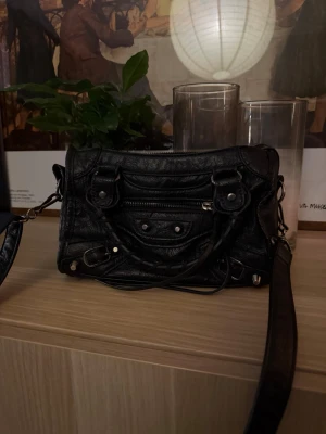 Svart handväska i läder - Cool svart handväska i  mjukt läder med klassiska nitar och dragkedjedetaljer. Väskan har både handtag och en lång axelrem, samt ett rymligt huvudfack och en mindre ficka framtill. Perfekt för dig som gillar edgy och tidlös design.