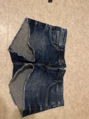 Mörkblå jeansshorts med rå kant - Snygga mörkblå jeansshorts med fransig, rå kant och klassisk femficksdesign. Shortsen har knapp och dragkedja framtill samt slitna detaljer för en cool look. Perfekta för sommaren och enkla att matcha med allt.