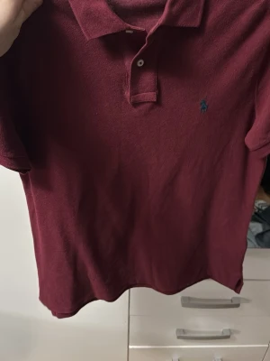 Vinröd pikétröja från Ralph Lauren - Säljer en vinröd pikétröja från Ralph Lauren med klassisk krage och två knappar framtill. Tröjan har korta ärmar och den ikoniska broderade loggan i blått på bröstet. Perfekt för en clean och stilren look.