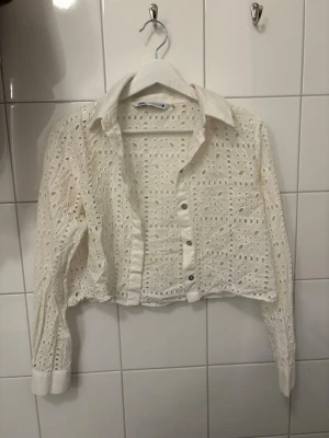 Vit croppad spetsblus från Zara - Säljer en vit croppad skjorta från Zara med genombruten spets och långa ärmar. Skjortan har klassisk krage, knappar framtill och ett luftigt, broderat mönster som ger en romantisk vibe.