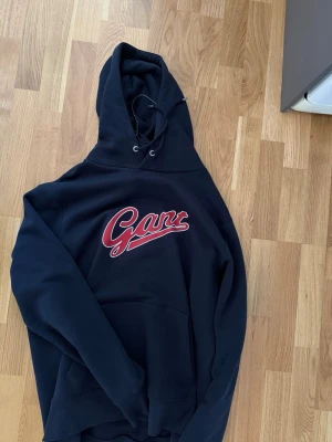Svart hoodie från Gant med tryck - Snygg svart hoodie från Gant med röd och vit logga tryckt på bröstet. Klassisk modell med huva och dragsko samt en stor magficka framtill. Perfekt för en avslappnad och trendig stil. Provad men aldrig använd.