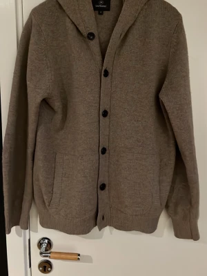 Beige kofta med huva från InWear - Mysig beige kofta från InWear med huva och knäppning framtill. Koftan har två fickor, ribbade muddar och en avslappnad passform. Perfekt för lager-på-lager och chill dagar.