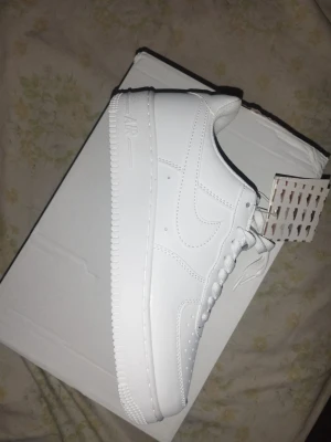 Nike Air Force 1 vita sneakers - Klassiska Nike Air Force 1 sneakers i helvitt läder med perforerad tå, vit sula och vita skosnören. Ikonisk Swoosh-logga på sidan och metallbricka med 'AF1' på snörningen. Perfekt för dig som gillar stilrena och tidlösa sneakers.