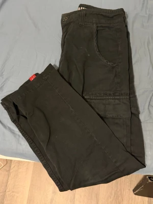 Svarta cargobyxor Alpha Industries - Säljer ett par svarta cargobyxor från Alpha Industries med klassiska cargofickor på sidorna och detaljer i orange på insidan av linningen. Byxorna har en avslappnad passform och är tillverkade i slitstarkt bomullstyg. Perfekta för en streetwear-look.