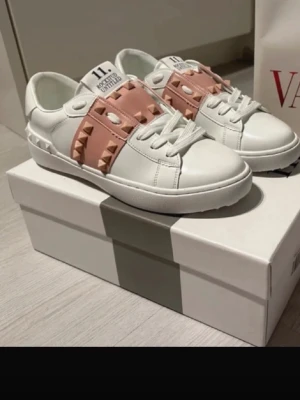 Valentino sneakers med rosa nitar - Snygga vita sneakers från Valentino med breda rosa band och coola nitar över vristen. Skorna har klassisk snörning och är tillverkade i skinn för en lyxig känsla. Perfekta för dig som vill sticka ut med en trendig och exklusiv look.
