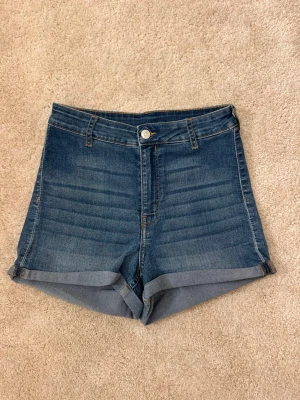 Blå denimshorts med uppvik - Snygga blå denimshorts med hög midja och uppvikta ben. Klassisk femficksdesign med knapp och dragkedja framtill. 