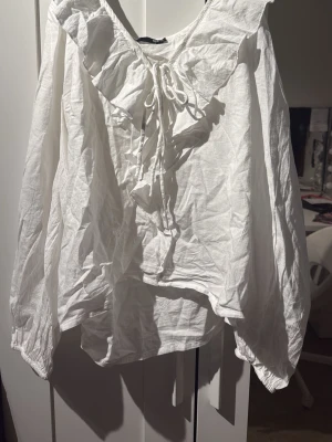 Vit blus med volang och knytband - Snygg vit blus i luftig bomull med volangdetalj längs ringningen och knytband framtill. Blusen har långa ärmar och en avslappnad passform som ger en romantisk vibe. Perfekt att styla med jeans eller kjol för en trendig look.