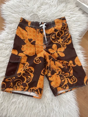 Bruna blommiga badshorts med snörning - Säljer ett par bruna badshorts med stora orange blommor och snörning i midjan. Shortsen har en praktisk sidoficka med knapp och vita kontrastsömmar. Perfekta för stranden eller poolen, riktigt sköna och snygga att bära!