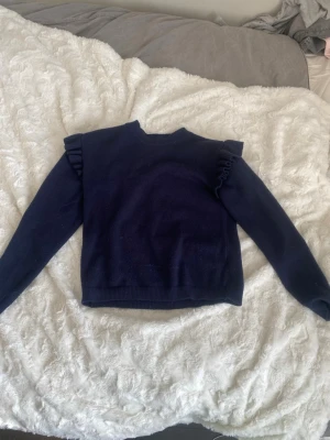 Mörkblå stickad tröja med volang - Mörkblå stickad tröja med långa ärmar och volangdetaljer på axlarna. Tröjan har rund halsringning och ribbstickade muddar vid ärmslut och nederkant. Perfekt för dig som gillar en enkel men ändå trendig look.