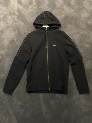 Svart hoodie med dragkedja  - Svart hoodie med hel dragkedja och huva. Gula detaljer längs dragkedjan och sömmarna på axlarna. Liten gul logga på bröstet. Perfekt för en chill och stilren look.