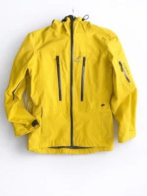 Gul vindjacka från Revolutionrace  - Snygg gul vindjacka från Revolutionrace med huva, svarta dragkedjor och flera praktiska fickor framtill och på ärmen. Jackan har justerbar huva och är tillverkad i slitstarkt material, perfekt för blåsiga dagar och utomhusaktiviteter.