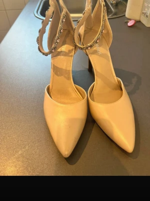 Beige pumps med strassdetaljer - Snygga beige pumps med spetsig tå och höga klackar. Skorna har en elegant rem runt vristen dekorerad med glittrande strass. Perfekta för dig som vill sticka ut med en stilren och trendig look. Dragkedja bak för enkel påtagning.