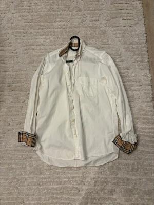 Vit Burberry skjorta  - Klassisk vit skjorta från Burberry med ikoniska beige-rutiga detaljer på krage och manschetter. Skjortan har bröstficka, knappar framtill och lång ärm. Perfekt för dig som gillar stilrena plagg med en twist.