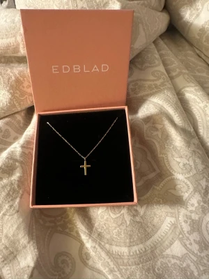 Guldfärgat korshalsband från Edblad - Snyggt halsband från Edblad med ett litet kors i guldfärg. Kedjan är tunn och elegant, och smycket levereras i original ask. Perfekt för dig som gillar stilrena accessoarer med en klassisk touch. (Aldrig använt)