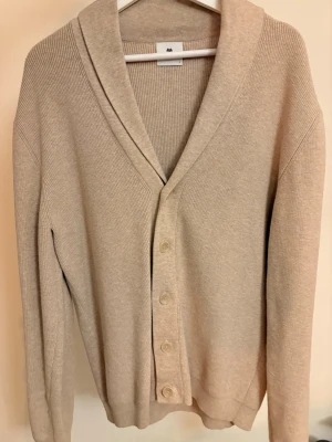 Beige kofta från John Henric - Stilren beige kofta från John Henric med sjalkrage och knäppning framtill. Tillverkad i mjukt, ribbstickat material som ger en klassisk och avslappnad look. Perfekt att slänga på sig över en t-shirt eller skjorta.