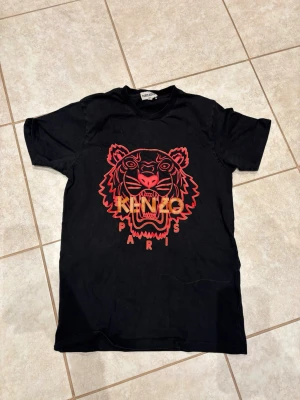Svart Kenzo t-shirt med tigertryck -  svart t-shirt från Kenzo med ett stort tigertryck i rött och orange på bröstet samt texten 'Kenzo Paris'. Klassisk passform och rund halsringning. 