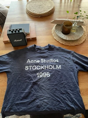 Blå Acne Studios t-shirt Stockholm - Snygg mörkblå t-shirt från Acne Studios med tryck 'STOCKHOLM 1996' på bröstet. Klassisk passform med rund hals och korta ärmar. Perfekt för dig som gillar stilrena och trendiga plagg med streetkänsla.