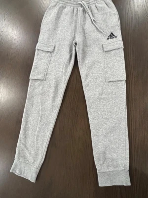 Grå cargobyxor från Adidas - Storlek är S junior. Snygga grå cargobyxor från Adidas med två stora fickor på sidorna och resår i midjan samt benslut. Byxorna har en avslappnad passform och är tillverkade i mjukt bomullsmaterial. Perfekta för en sportig och chill stil.
