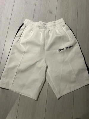 Vita Palm Angels shorts med svarta detaljer - Snygga vita shorts från Palm Angels med svarta sidor och broderad logga på vänster ben. De har elastisk midja och sportig look, perfekta för en avslappnad streetwear-stil.