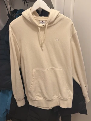 Beige hoodie från Champion - Säljer en stilren beige hoodie från Champion med broderad logga på bröstet. Hoodien har huva med dragsko och en stor magficka. Tillverkad i mjukt bomullsmaterial och har ribbade muddar vid ärmslut och nederkant. Perfekt för en avslappnad look. Passar M också 