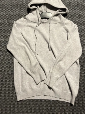Merinoulls tröja - En stilren ljusgrå hoodie med huva och dragsnören. Tröjan har långa ärmar, ribbade muddar och en avslappnad passform. Perfekt för en chill och enkel look. Materialet känns mjukt och bekvämt, passar till det mesta i garderoben.