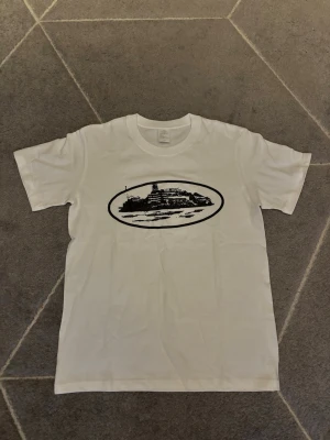 Vit t-shirt med svart Corteiz tryck - Vit t-shirt från Corteiz med ett svart tryck av ett landskap eller byggnad i oval form på bröstet. Klassisk passform och rund hals. Perfekt för en avslappnad stil.