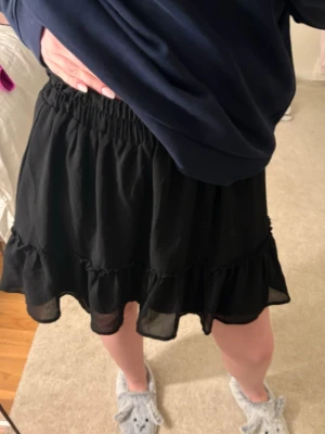 Svart volangkjol med resårmidja - Säljer en svart kortkjol med volangkant och resår i midjan. Kjolen har en luftig och flowy känsla, perfekt för att styla med en hoodie eller crop top. Materialet känns lätt och bekvämt.